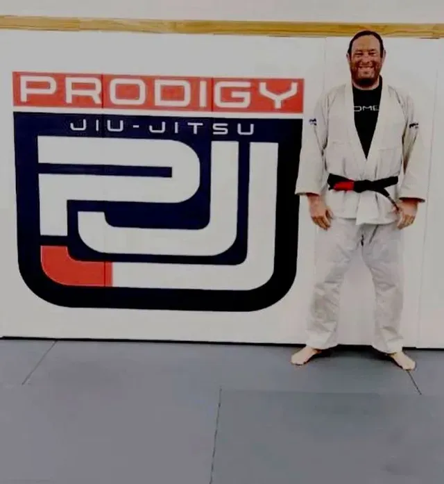 Coach Jason Zicherman in white gi with black belt in front of Prodigy Jiu-Jitsu logo