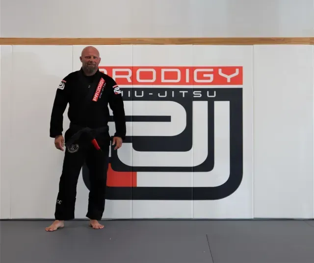 Coach Luke in black gi with belt in front of Prodigy Jiu-Jitsu logo