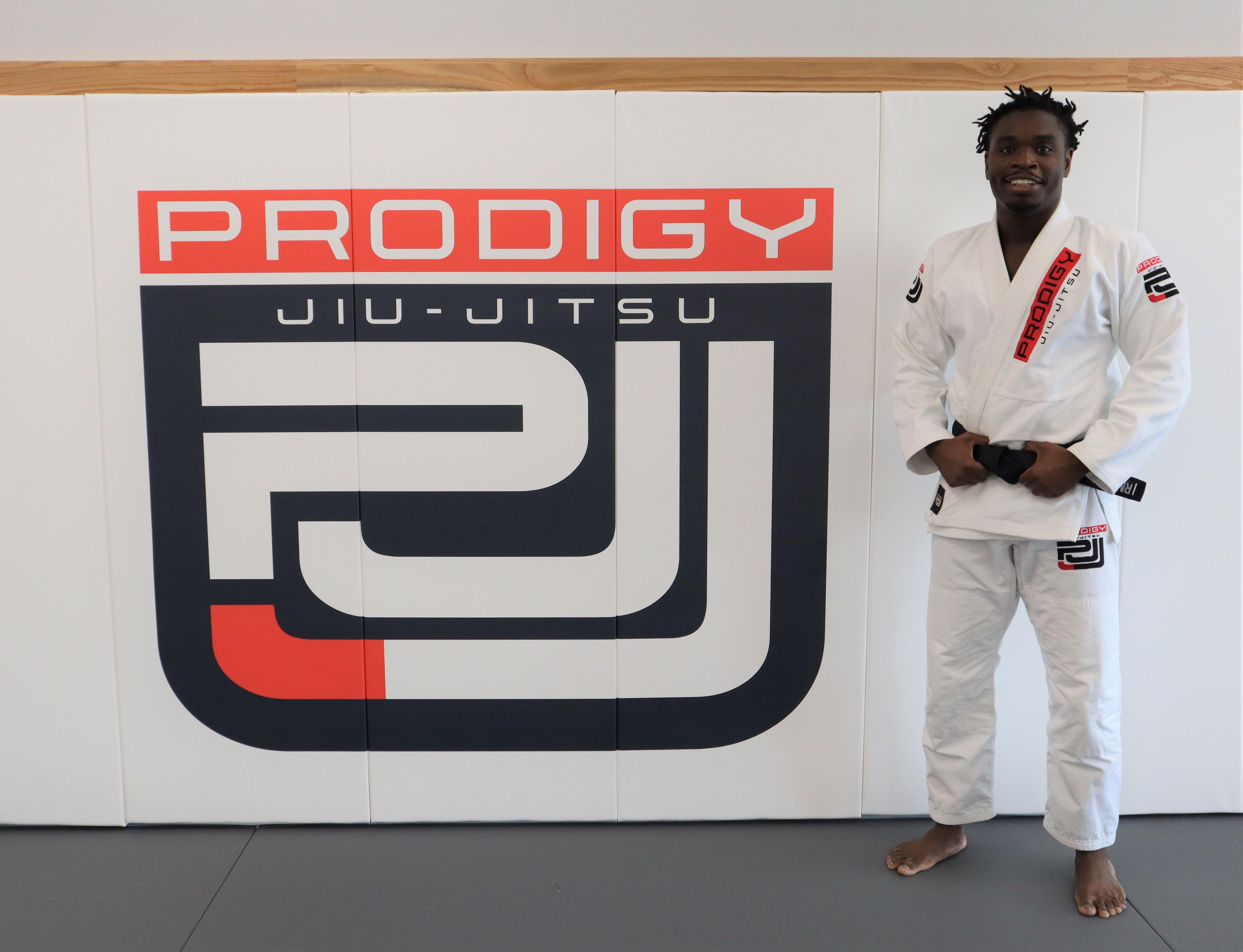 Coach Shakiyl Jones in white gi with black belt standing in front of Prodigy Jiu-Jitsu logo