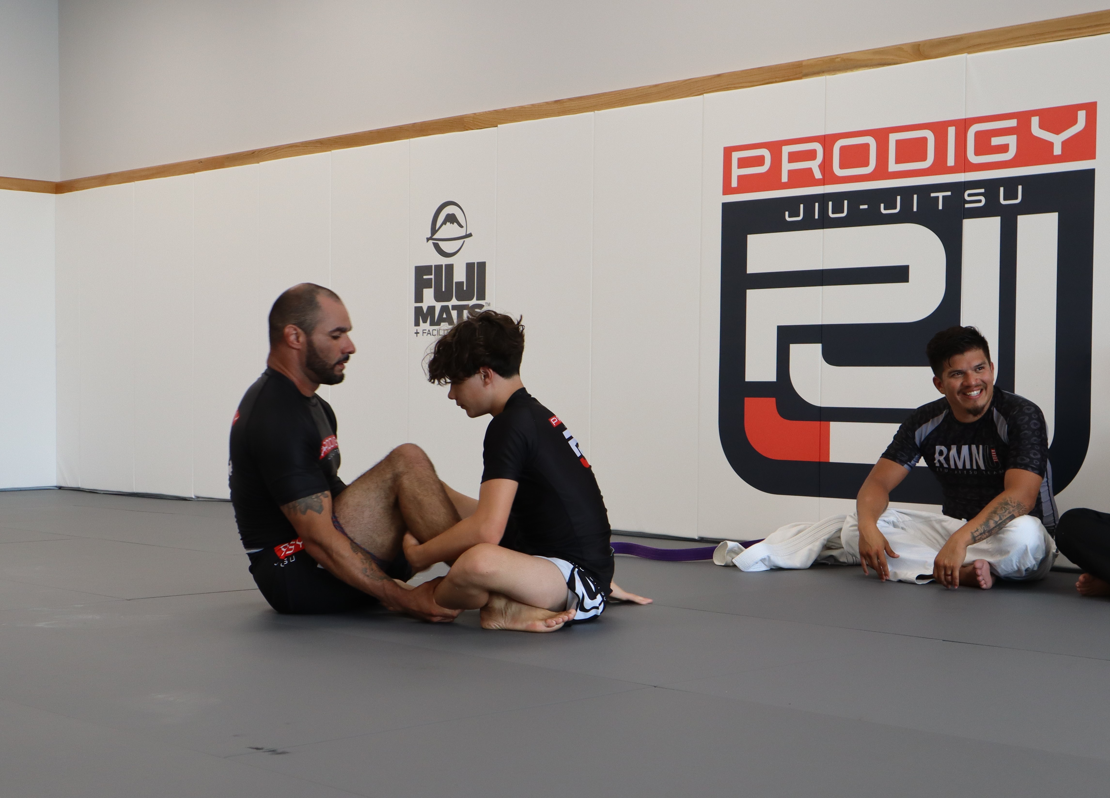 Prodigy Jiu-Jitsu coach giving one-on-one instruction to students on the mat in front of the academy logo